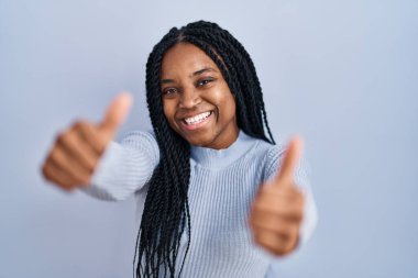 African american woman standing over blue background approving doing positive gesture with hand, thumbs up smiling and happy for success. winner gesture. 