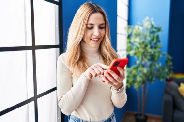 Young blonde woman using smartphone standing at home