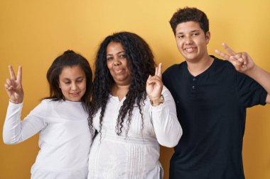 Anne, kız ve oğulun ailesi sarı arka planda durup kameraya bakıp zafer işareti yaparken gülümsüyor. İki numara.. 