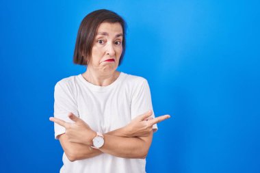 Middle age hispanic woman standing over blue background pointing to both sides with fingers, different direction disagree 