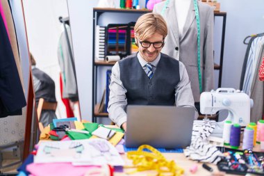 Young blond man tailor smiling confident using laptop at clothing factory