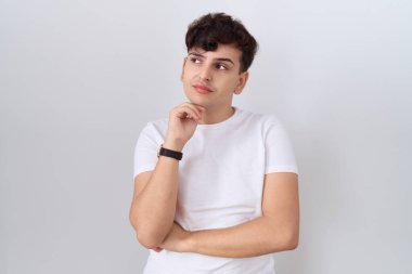 Gündelik beyaz tişört giyen, eli çenesinde soru soran, düşünceli bir genç. Düşünceli bir yüzle gülümsüyor. şüphe kavramı. 