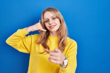 Young caucasian woman standing over blue background smiling doing talking on the telephone gesture and pointing to you. call me. 