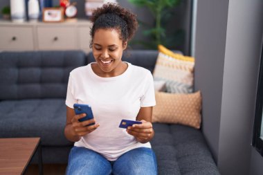 Akıllı telefon ve kredi kartı kullanan Afrikalı Amerikalı bir kadın evdeki kanepede oturuyor.
