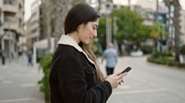 Genç, güzel, İspanyol bir kadın sokakta akıllı telefon kullanarak gülümsüyor.
