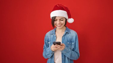 Kafkasyalı genç bir kadın noel şapkası takıyor ve izole edilmiş kırmızı arka plan üzerinde akıllı telefon kullanıyor.