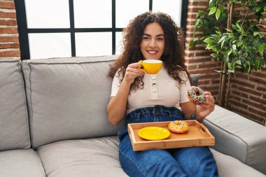 Genç, güzel, İspanyol bir kadın kahvaltısını evde, koltukta yapıyor.