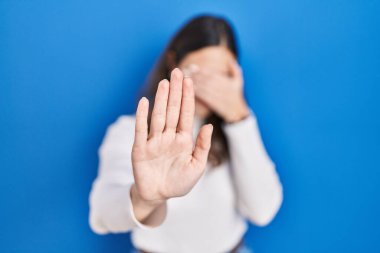 Mavi arka planda duran genç İspanyol kadın elleriyle gözlerini kapatıyor ve üzgün ve korku ifadesiyle dur hareketi yapıyor. utangaç ve olumsuz kavram. 
