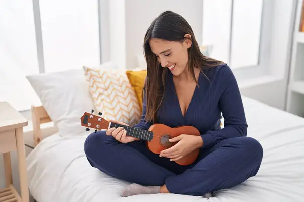 Young beautiful hispanic woman playing ukulele sitting on bed at bedroom