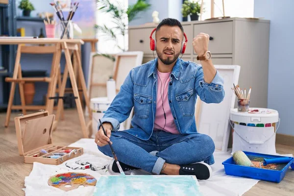 Young hispanic man painter sitting on the floor at art studio annoyed and frustrated shouting with anger, yelling crazy with anger and hand raised