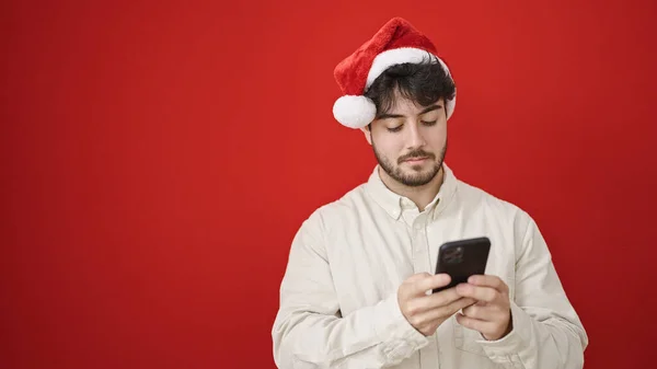 Νεαρός Ισπανός Που Φοράει Χριστουγεννιάτικο Καπέλο Χρησιμοποιώντας Smartphone Απομονωμένο Κόκκινο — Φωτογραφία Αρχείου