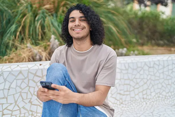 Young latin man using smartphone sitting on bench at park