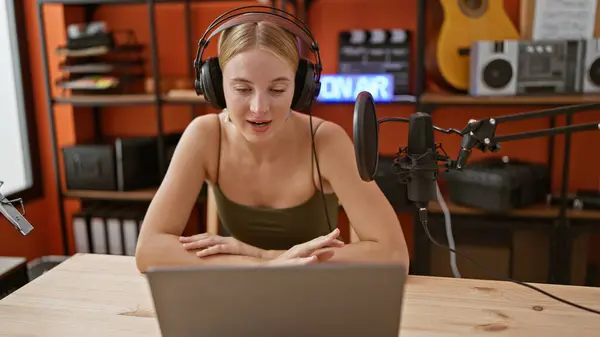 Young blonde woman radio reporter speaking using headphones and laptop at podcast studio