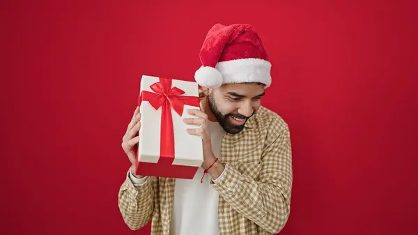 stock image Young hispanic man smiling confident wearing christmas hat hearing gift sound over isolated red background