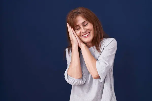 Middle age woman standing over blue background sleeping tired dreaming and posing with hands together while smiling with closed eyes.