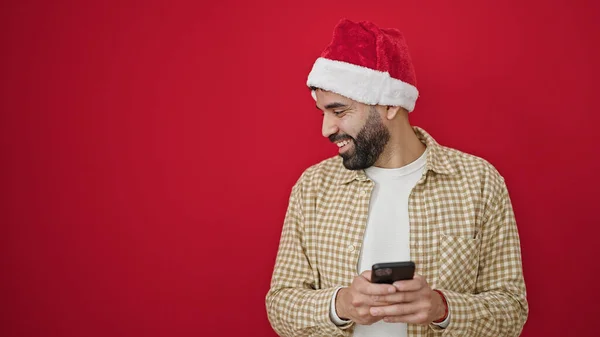 Junger Hispanischer Mann Trägt Weihnachtsmütze Mit Smartphone Vor Isoliertem Roten — Stockfoto