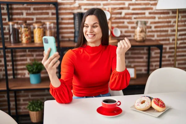 Young hispanic woman eating doughnuts and drinking a cup of coffee doing video call screaming proud, celebrating victory and success very excited with raised arm