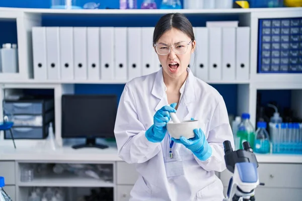 Chinese young woman working at scientist laboratory mixing angry and mad screaming frustrated and furious, shouting with anger. rage and aggressive concept.