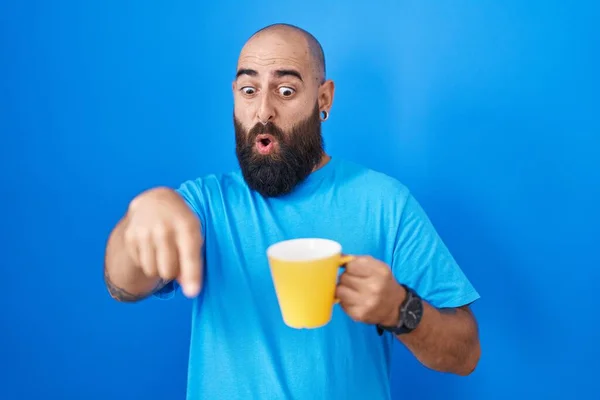 Young hispanic man with beard and tattoos drinking a cup of coffee pointing down with fingers showing advertisement, surprised face and open mouth