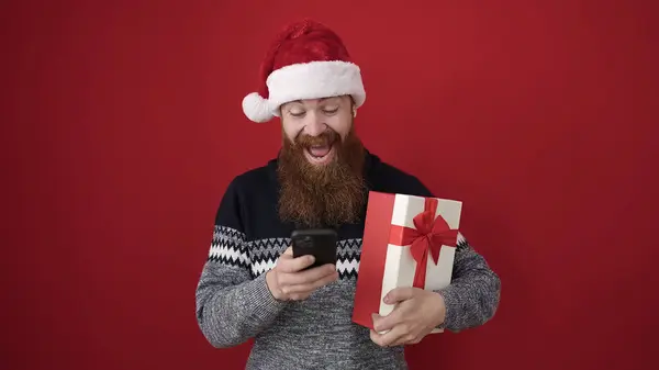 Junger Rothaariger Mann Hält Weihnachtsgeschenk Mit Smartphone Über Isoliertem Rotem — Stockfoto