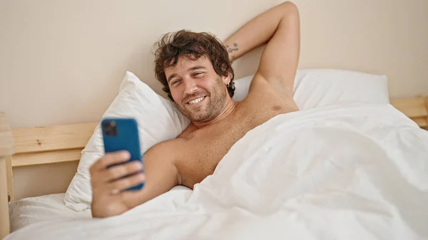 年轻的他惊慌失措的男人赤身裸体躺在床上 用智能手机在卧室里自作主张 — 图库照片