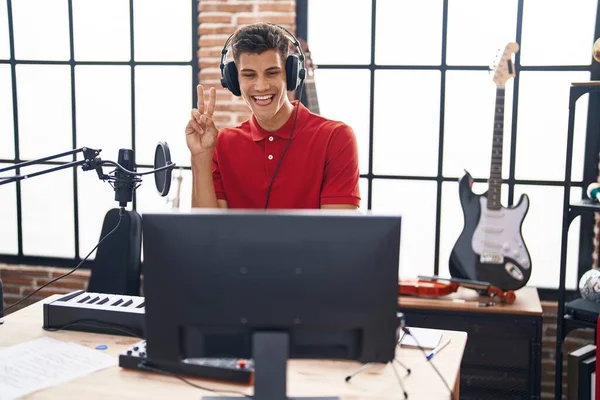 Young hispanic man playing piano keyboard at music studio smiling with happy face winking at the camera doing victory sign. number two.
