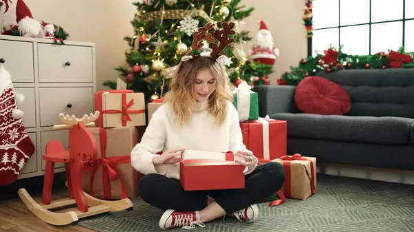 年轻的金发女子打开包装 坐在自家圣诞树旁的地板上 — 图库照片