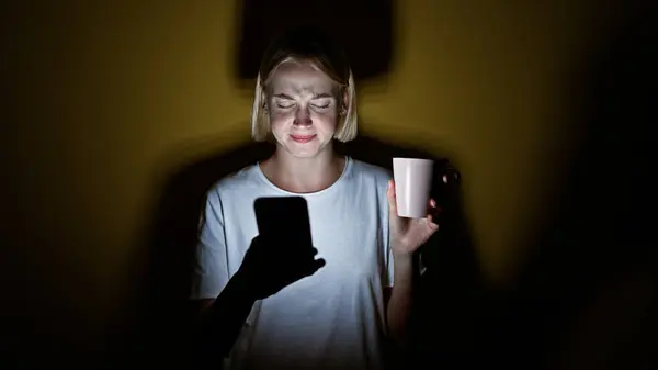 Jovem Loira Usando Smartphone Segurando Xícara Café Cansado Sobre Fundo — Fotografia de Stock