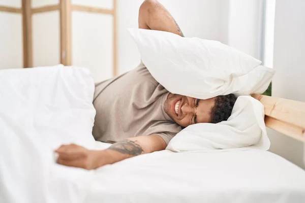 African american man suffering for noise covering ears with pillow at bedroom