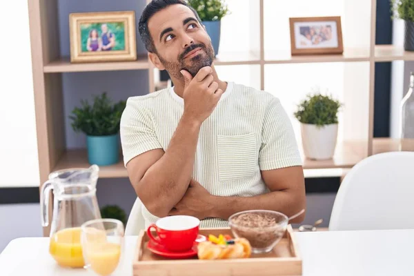 Hispanic man with beard eating breakfast smiling looking confident at the camera with crossed arms and hand on chin. thinking positive.