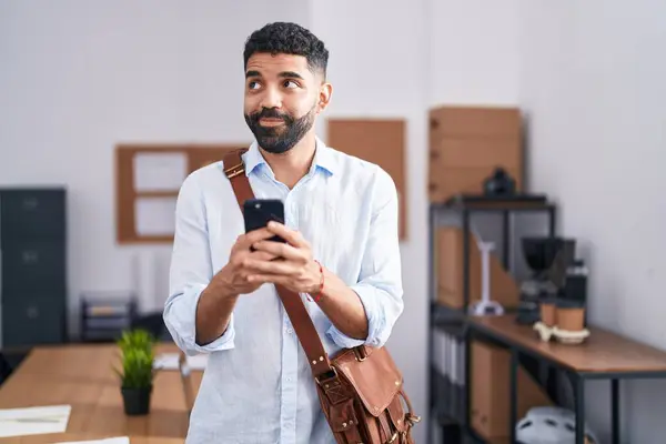 一个留着胡子的西班牙裔男人在办公室里用智能手机微笑着看着旁边 目不转睛地看着外面想 — 图库照片