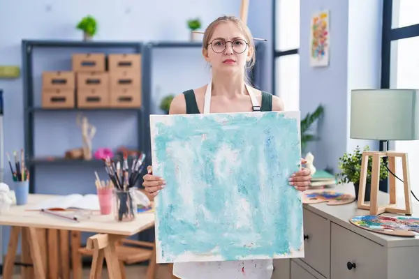 年轻的高加索女人在艺术工作室拿着画布 充满怀疑和紧张 因为问题皱着眉头 消极的人 — 图库照片