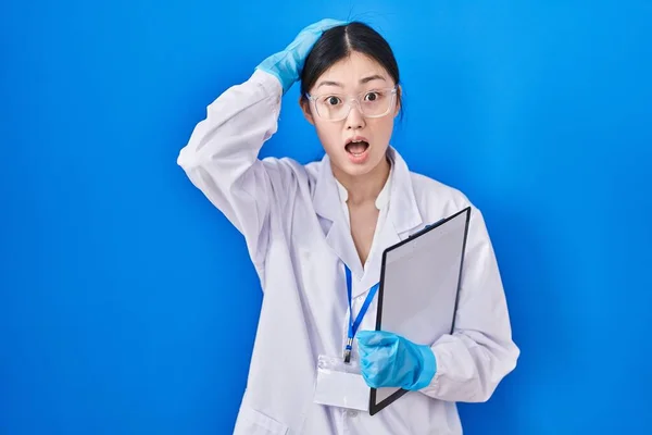 Chinese young woman working at scientist laboratory crazy and scared with hands on head, afraid and surprised of shock with open mouth