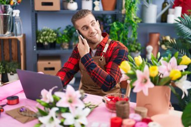 Young caucasian man florist talking on smartphone using laptop at flower shop
