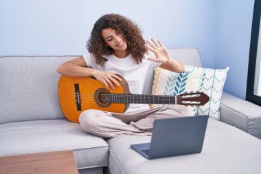 Young beautiful hispanic woman having online classical guitar class sitting on sofa at home