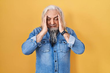 Sarı arka planda duran gri saçlı orta yaşlı bir adam elleri başının üstünde, stres yüzünden başı ağrıyor. migren ağrısı. 