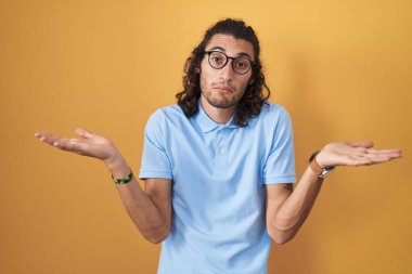 Young hispanic man standing over yellow background clueless and confused expression with arms and hands raised. doubt concept. 