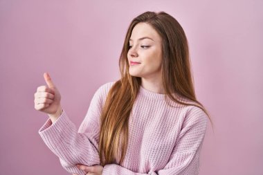 Pembe arka planda duran beyaz bir kadın gururlu görünüyor, gülümsüyor. Kenarda baş parmak hareketi yapıyor. 
