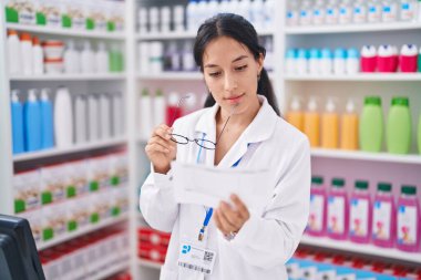 Genç ve güzel İspanyol kadın eczacı elinde gözlükle eczanede reçete okuyor.