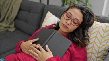 Genç, güzel, İspanyol bir kadın elinde kitapla kanepede uzanıp evde uyuyor.