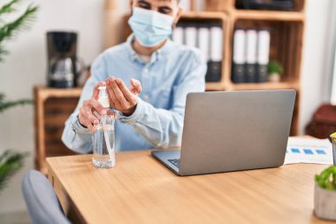 Young hispanic man business worker wearing medical mask using sanitizer gel hands at office