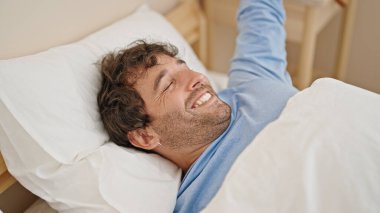 Young hispanic man waking up stretching arms at bedroom