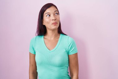 Young hispanic woman standing over pink background smiling looking to the side and staring away thinking. 