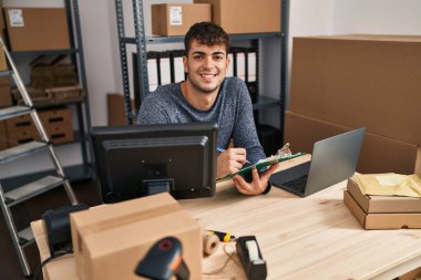 Young hispanic man ecommerce business worker using laptop writing on document at office
