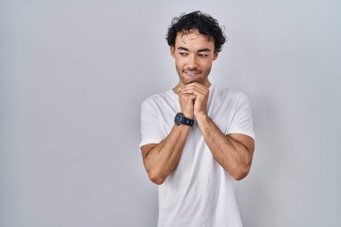 Hispanik adam arka planda dikiliyor. Gergin ve heyecanlı. Elleri çenesinde yan tarafa bakıyor. 