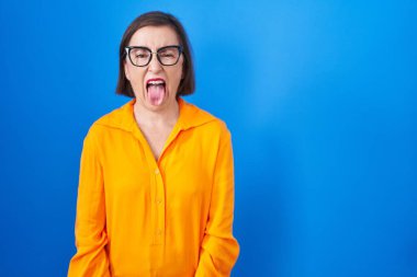 Mavi arka planda duran gözlüklü, orta yaşlı İspanyol kadın komik bir ifadeyle dilini dışarı çıkartıyor. duygu kavramı. 