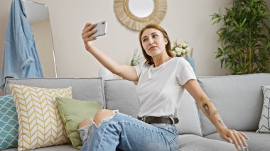 Evdeki kanepede oturmuş akıllı telefonuyla selfie çeken genç bir kadın.