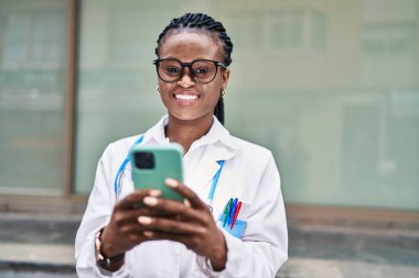 Afro-Amerikalı kadın doktor hastanede akıllı telefon kullanarak gülümsüyor.