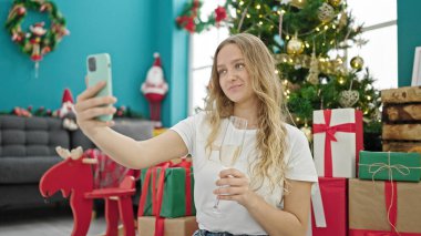 Genç sarışın kadın Noel 'i kutluyor. Akıllı telefonuyla selfie çekiyor. Evde şampanya içiyor.