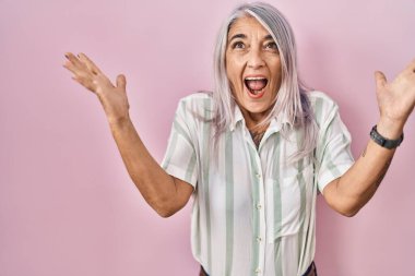 Pembe arka planda duran gri saçlı orta yaşlı kadın zaferi mutlu bir gülümsemeyle ve ellerini kaldırarak kazanan ifadeyle kutluyor. 